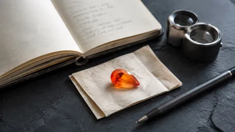 A pear-cut fire opal on gemstone paper beside a product notebook, jeweler's loupe, and pen on a slate work surface.